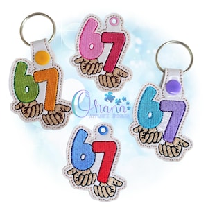 May include: Keychains with the number 67 in various colors, including green, orange, pink, red, blue, and purple. Each keychain features the number held by a pair of hands. The keychains have a white leather strap and a metal ring.