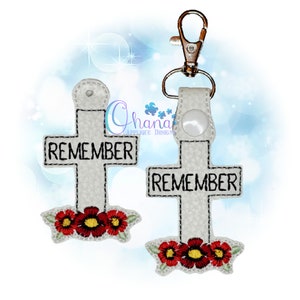 May include: White cross keychain with the word "REMEMBER" on it. The cross is decorated with three red flowers at the bottom.