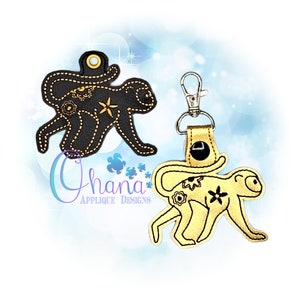 Może przedstawiać: Dwa haftowane breloczki z małpą, jeden czarny, a drugi złoty, z kwiatowymi wzorami. Czarny brelok z małpą ma pętlę do przymocowania do torby lub paska. Złoty brelok z małpą ma metalową klamrę do przymocowania do kluczy.