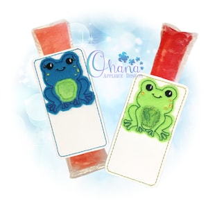 May include: Two white fabric pouches with a frog applique on each. The frog on the left pouch is blue with a green belly and the frog on the right pouch is green with a green belly. The pouches are designed to hold popsicles.