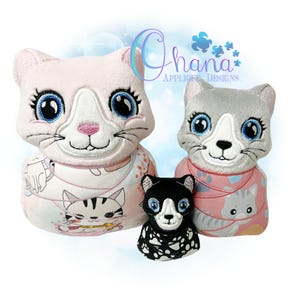 May include: Three plush cat toys in different sizes. The largest cat is pink with white accents and blue eyes. The middle cat is gray with white accents and blue eyes. The smallest cat is black with white accents and blue eyes. All three cats have embroidered facial features.