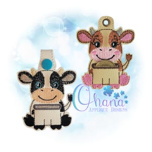 Può includere: Due portachiavi di mucca ricamati con un porta monete. La mucca a sinistra è bianca e nera con un bottone blu. La mucca a destra è marrone e bianca con zoccoli rosa e un porta monete marrone.
