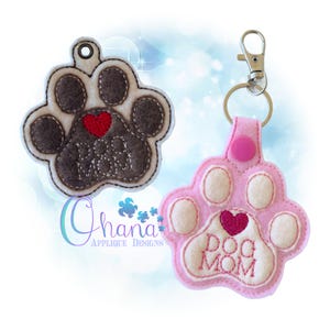 May include: Two felt paw print keychains. One is brown with a red heart and the text "Dog Mom". The other is pink with a pink heart and the text "Dog Mom".