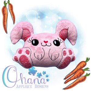May include: Pink plush bunny with embroidered details, including eyes, nose, and paws. The bunny is sitting with its paws in front of it. There are carrots in the background.