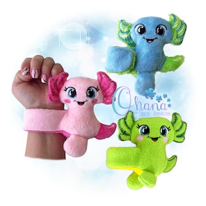 May include: Three plush axolotl toys in pink, blue, and green. Each has a smiling face, large eyes, and frilly gills. One is attached to a pink wristband. The toys are soft and colorful, perfect for children.