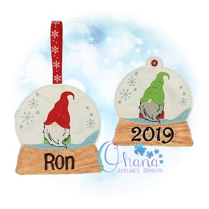 Könnte beinhalten: Zwei gestickte Weihnachtsornamente mit Gnomen in Schneekugeln. Das erste Ornament hat einen Gnom mit roter Mütze und den Namen "Ron" auf der Basis. Das zweite Ornament hat einen Gnom mit grüner Mütze und das Jahr "2019" auf der Basis.