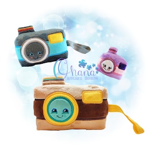 May include: Three plush toy cameras with smiling faces. The cameras are brown, blue, and purple. The brown camera has a yellow strap. The blue camera has a gray strap. The purple camera has a pink strap. The cameras are against a blue and white background.