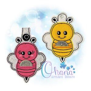 May include: Two felt bee-shaped coin purses. One is pink with silver glitter wings, the other is yellow with white wings. Each has a slot for coins and a smiling face. The text "Ohana Applique Designs" is visible.