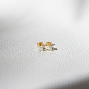 May include: A pair of gold earrings with light blue square-shaped gemstones. The earrings are on a white surface.