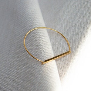 May include: A gold-toned bangle bracelet with a straight bar design. The bracelet is on a white fabric background.