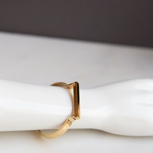 Op de afbeelding: Een gouden armband met een rechthoekig ontwerp. De armband is open en heeft een scharnier sluiting.