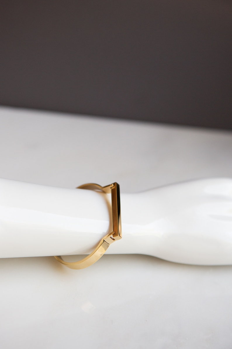 Op de afbeelding: Een gouden armband met een rechthoekig ontwerp. De armband is open en heeft een scharnier sluiting.