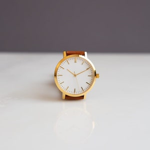 May include: A gold-toned wristwatch with a white face and gold hands. The watch has a brown leather band. The watch is set against a white and gray background.