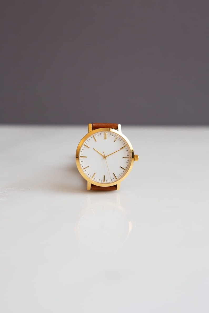 May include: A gold-toned wristwatch with a white face and gold hands. The watch has a brown leather band. The watch is set against a white and gray background.