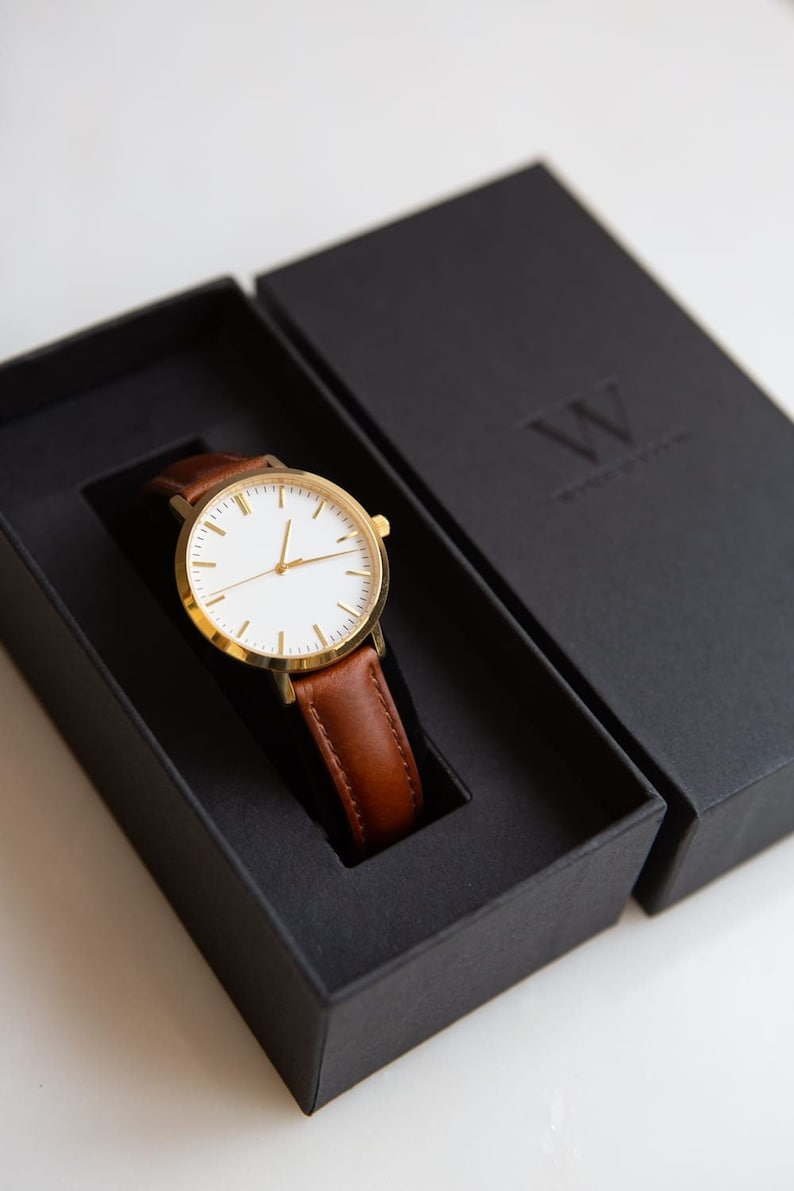 May include: A gold-tone wristwatch with a white face and brown leather band rests in a black box. The watch has a minimalist design. The box is open, revealing the watch. The watch is a classic accessory.