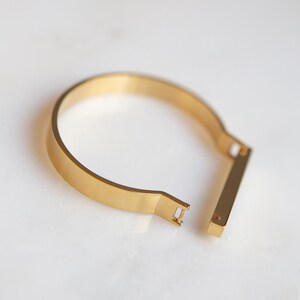 Op de afbeelding: Een gouden metalen armband met een eenvoudig, minimalistisch ontwerp. De armband heeft een plat, rechthoekig bandje dat open en dicht gaat met een schuifmechanisme.