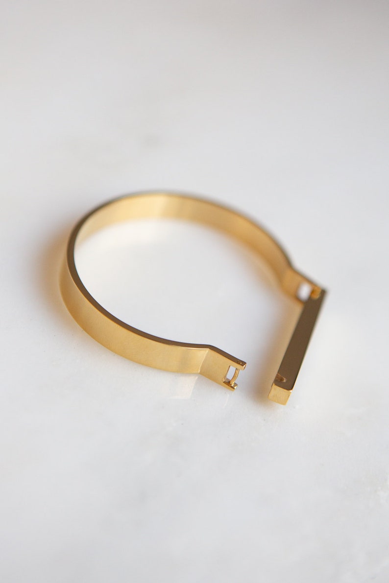 Op de afbeelding: Een gouden metalen armband met een eenvoudig, minimalistisch ontwerp. De armband heeft een plat, rechthoekig bandje dat open en dicht gaat met een schuifmechanisme.