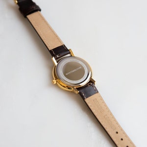 May include: A gold-tone watch with a brown leather band. The back of the watch case is engraved with the text "WYNTERSBLOOM".