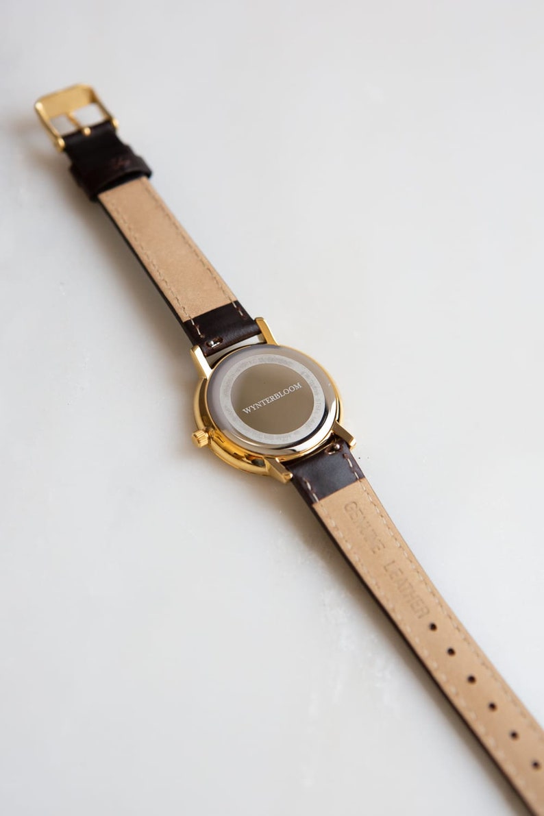 May include: A gold-tone watch with a brown leather band. The back of the watch case is engraved with the text "WYNTERSBLOOM".
