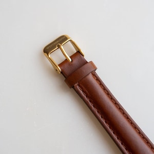 May include: Close-up of a brown leather watch strap with a gold-colored buckle. The buckle is rectangular and has the word "WINTERBLOOM" engraved on it. The leather strap is stitched along the edges.