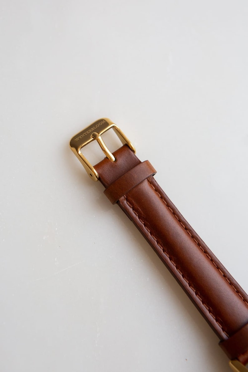 May include: Close-up of a brown leather watch strap with a gold-colored buckle. The buckle is rectangular and has the word "WINTERBLOOM" engraved on it. The leather strap is stitched along the edges.