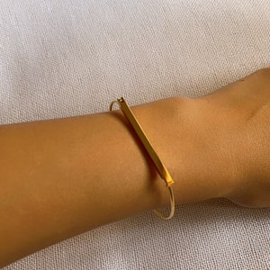 Może przedstawiać: Złota bransoletka typu bangle z prostym wzorem w postaci pręta. Bransoletka jest otwarta i ma prosty, minimalistyczny styl.