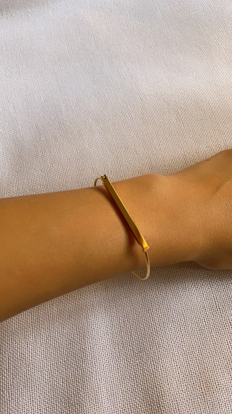 Może przedstawiać: Złota bransoletka typu bangle z prostym wzorem w postaci pręta. Bransoletka jest otwarta i ma prosty, minimalistyczny styl.