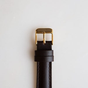 May include: A brown leather watch strap with a gold buckle. The buckle has the text "WYNTERSBLOOM" engraved on it.