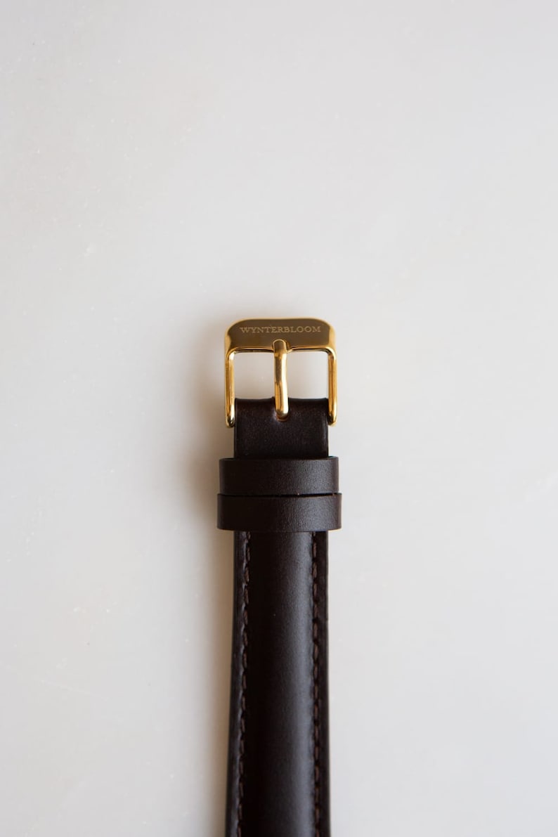 May include: A brown leather watch strap with a gold buckle. The buckle has the text "WYNTERSBLOOM" engraved on it.