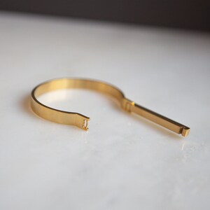 Op de afbeelding: Een gouden metalen armband met een uniek open ontwerp. De armband bestaat uit twee delen die met een eenvoudige sluiting worden verbonden.