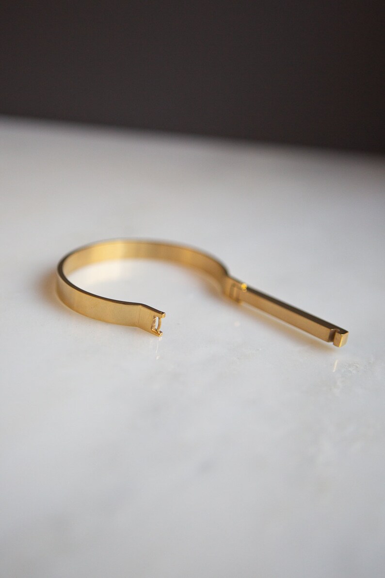 Op de afbeelding: Een gouden metalen armband met een uniek open ontwerp. De armband bestaat uit twee delen die met een eenvoudige sluiting worden verbonden.
