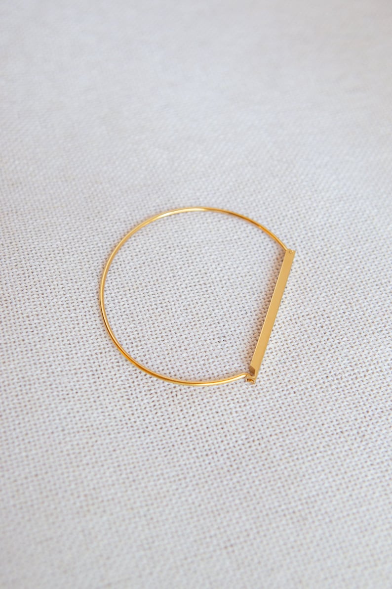 Może przedstawiać: Złota bransoletka typu bangle z prostym wzorem w postaci pręta. Bransoletka jest otwarta i ma prosty, minimalistyczny styl.