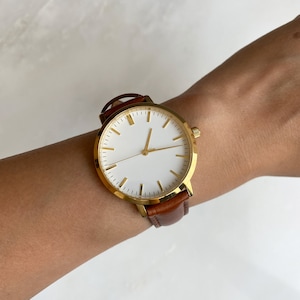 May include: A gold-toned wristwatch with a white face and gold markers. The watch has a brown leather band. The watch is on a wrist.