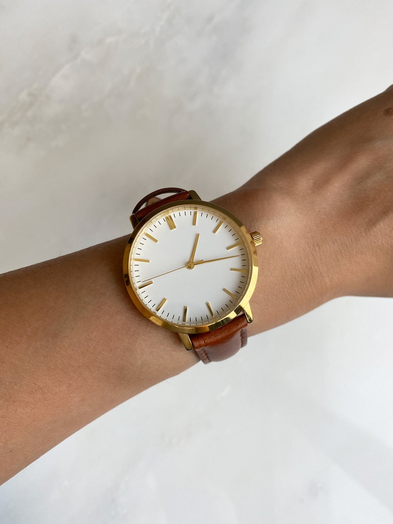 May include: A gold-toned wristwatch with a white face and gold markers. The watch has a brown leather band. The watch is on a wrist.