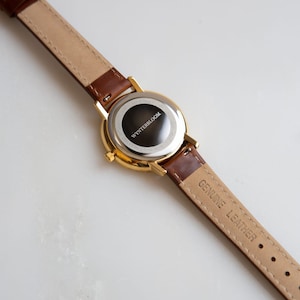 May include: A gold-toned wristwatch with a brown leather band. The watch face is black with the text "WYNTERBLOOM". The band is marked "GENUINE LEATHER". The watch is shown against a white background.