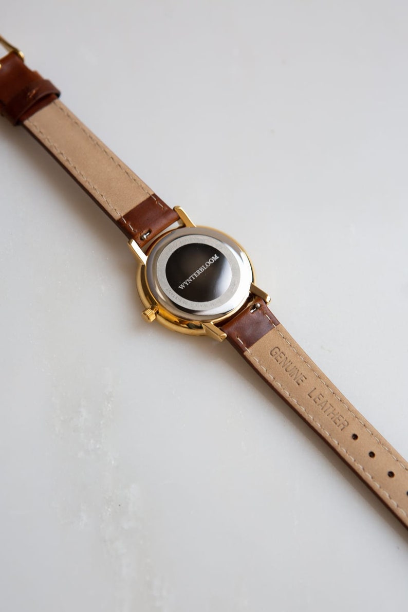 May include: A gold-toned wristwatch with a brown leather band. The watch face is black with the text "WYNTERBLOOM". The band is marked "GENUINE LEATHER". The watch is shown against a white background.