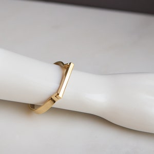 Op de afbeelding: Een gouden armband met een vierkante vorm en een plat, rechthoekig balkontwerp.
