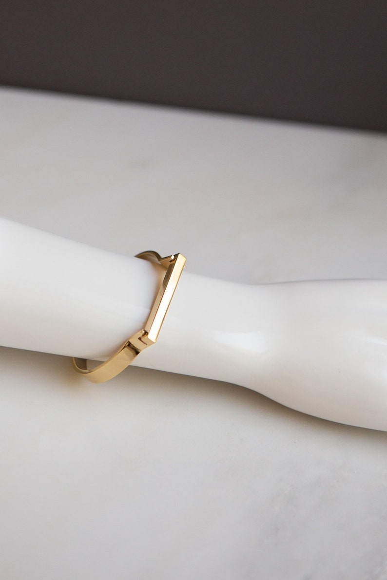 Op de afbeelding: Een gouden armband met een vierkante vorm en een plat, rechthoekig balkontwerp.