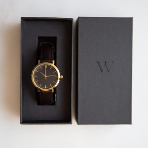 May include: A gold watch with a black face and brown leather band in a black box. The box has the text "WYTHENSHAWS" on it.