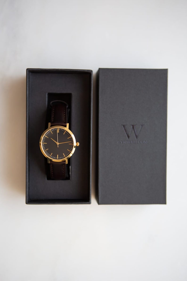 May include: A gold watch with a black face and brown leather band in a black box. The box has the text "WYTHENSHAWS" on it.