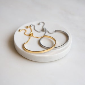 May include: Two minimalist bracelets, one gold and one silver, with adjustable sliding knots. The bracelets are on a white surface.