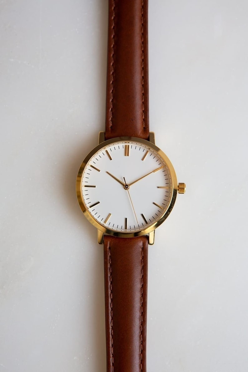 May include: A minimalist wristwatch with a gold-colored case and a white watch face. The watch has gold hands and markers, and a brown leather band. The watch is a classic design, suitable for everyday wear.