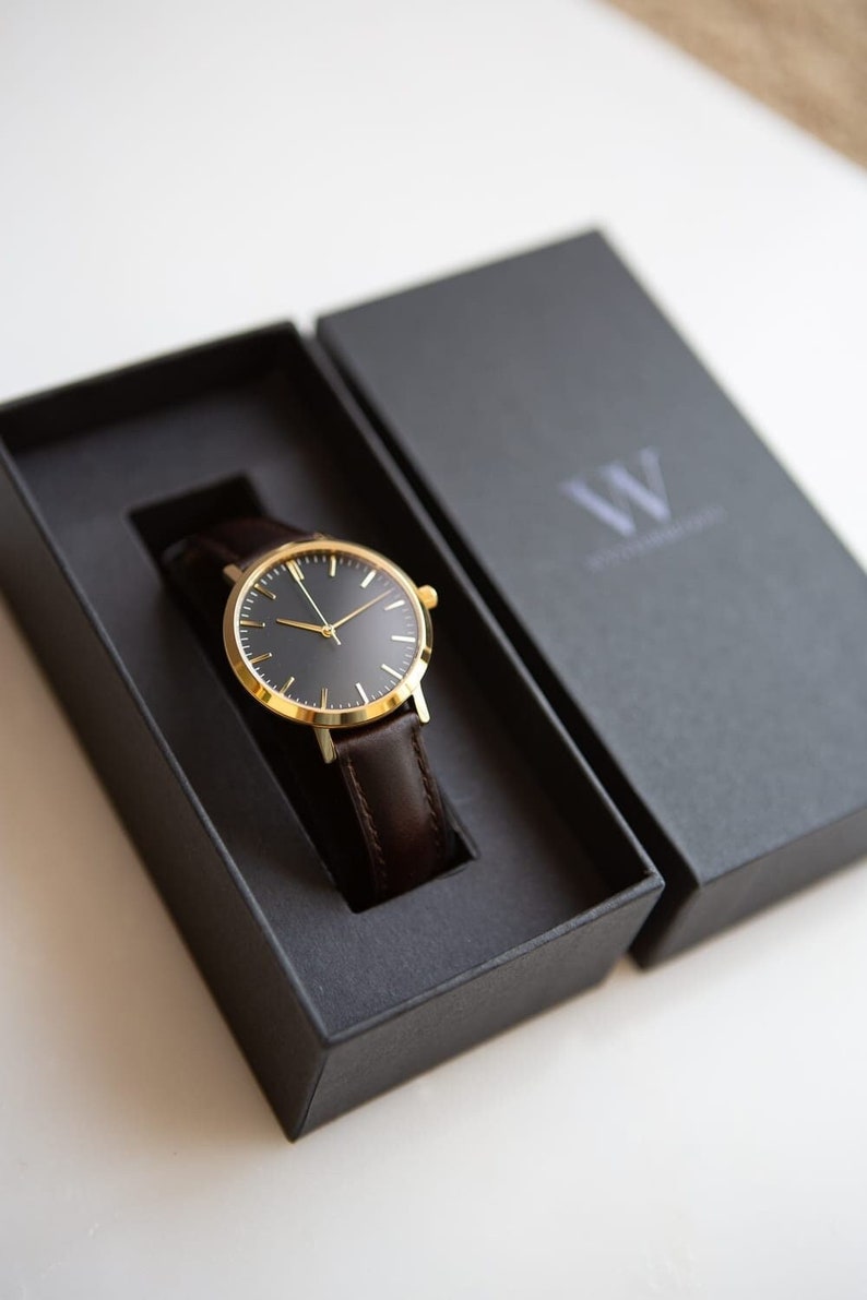 May include: A gold watch with a black face and brown leather band is displayed in a black box. The box has the letter "W" on the lid.