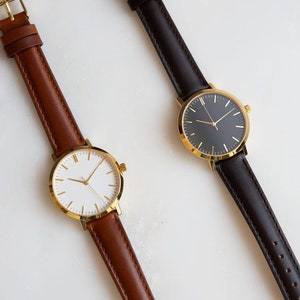 May include: Two wristwatches with gold-tone cases and different colored leather bands. The watch on the left has a white face and brown band, while the watch on the right has a black face and a black band.