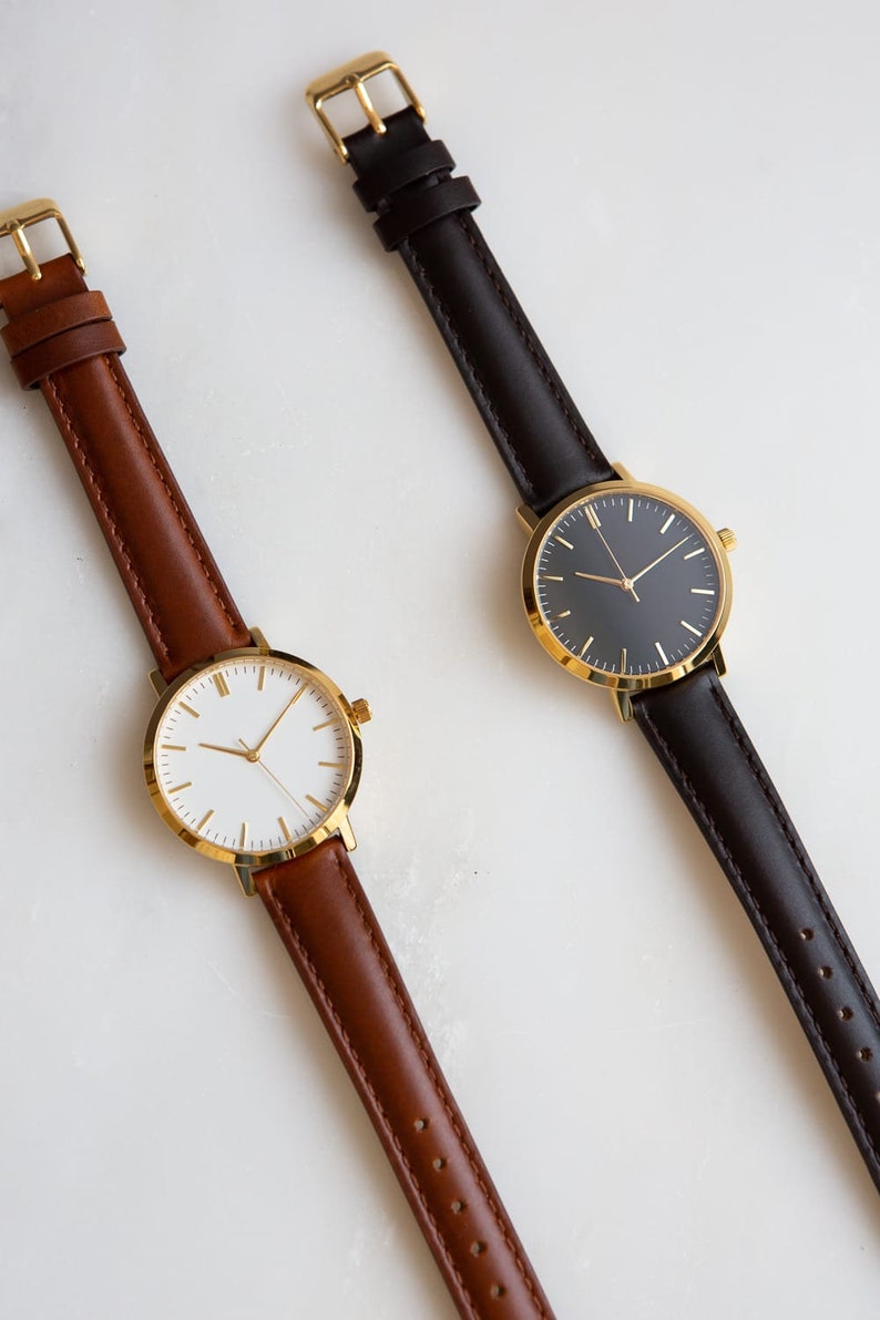 May include: Two wristwatches with gold-tone cases and different colored leather bands. The watch on the left has a white face and brown band, while the watch on the right has a black face and a black band.