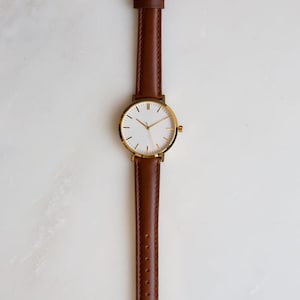 May include: A minimalist wristwatch with a gold-colored case and a brown leather strap. The watch face is white with gold-colored hands and markers. The strap is a rich brown color and has a gold-colored buckle. The watch is displayed on a white surface.