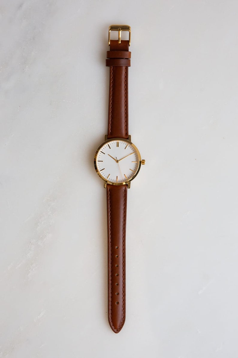 May include: A minimalist wristwatch with a gold-colored case and a brown leather strap. The watch face is white with gold-colored hands and markers. The strap is a rich brown color and has a gold-colored buckle. The watch is displayed on a white surface.