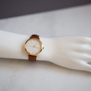 May include: A gold-tone wristwatch with a white face and brown leather band.