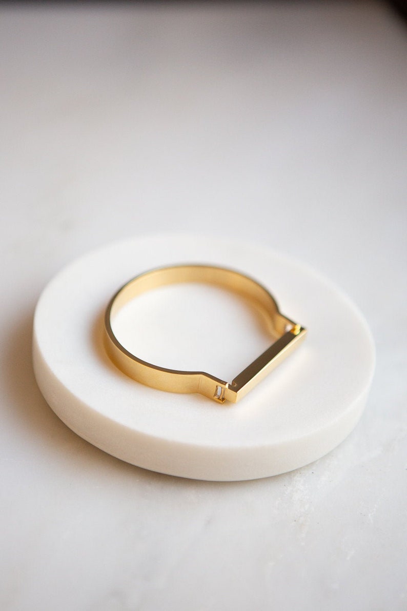 Op de afbeelding: Een gouden armband met een stangensluiting. De armband ligt op een wit marmeren oppervlak.