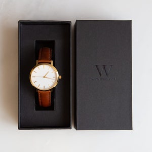 May include: A gold-tone wristwatch with a white face and brown leather band is displayed in an open black box. The watch is presented next to a closed black box with the embossed text "WYNTERBLOOM". The watch has a classic, minimalist design.